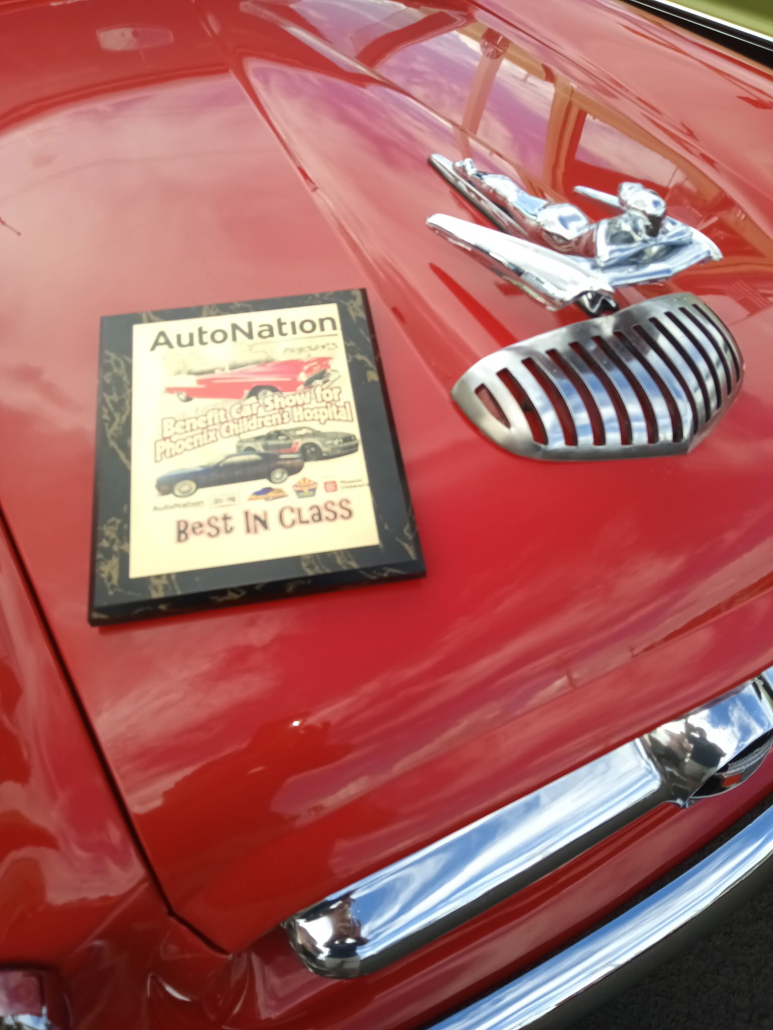 Red vintage car with AutoNation Best in Class award plaque and chrome hood ornament detail.