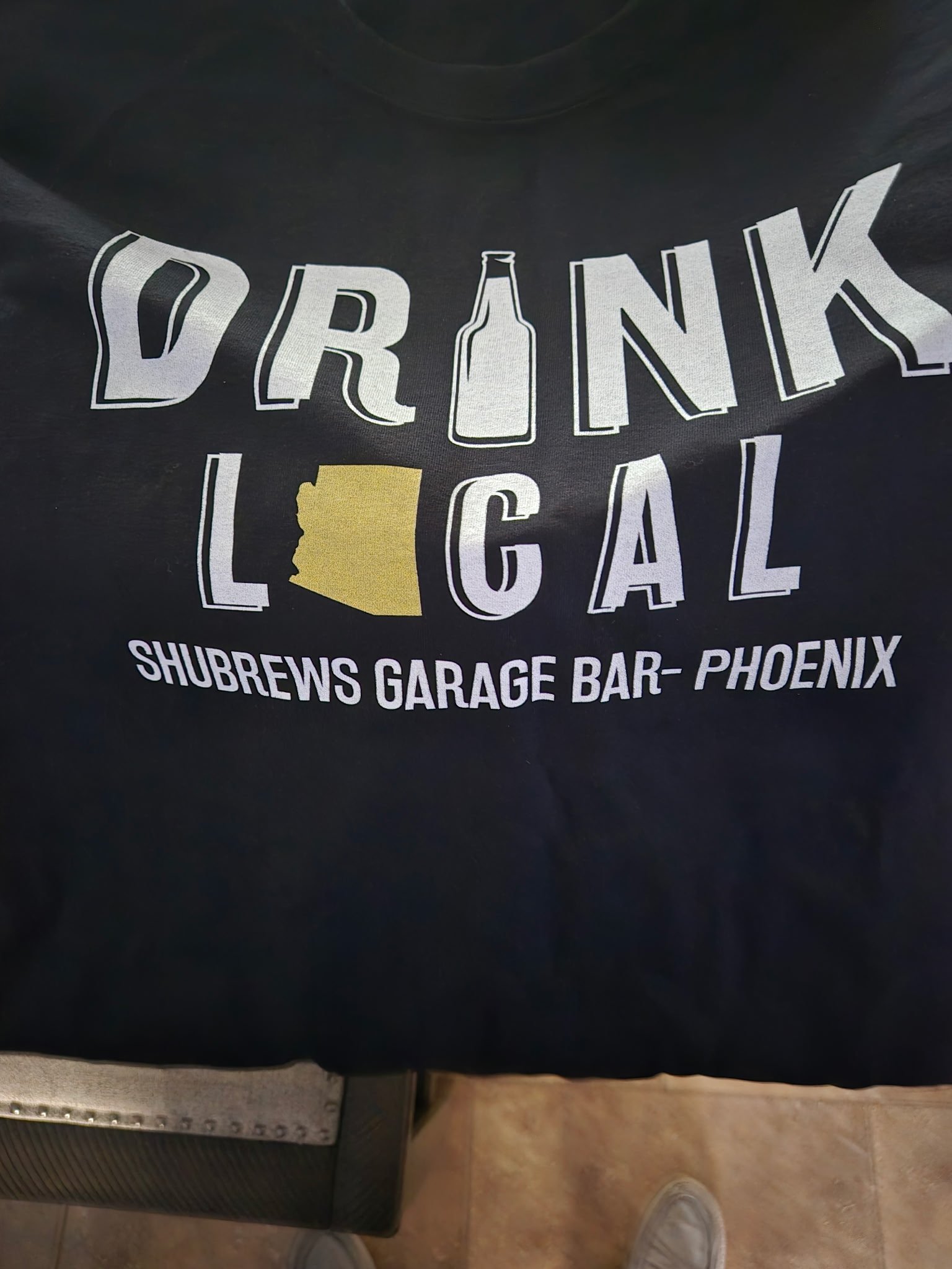 Black t-shirt with "Drink Local" text, beer bottle icon, and gold Arizona silhouette for ShuBrews Garage Bar Phoenix.