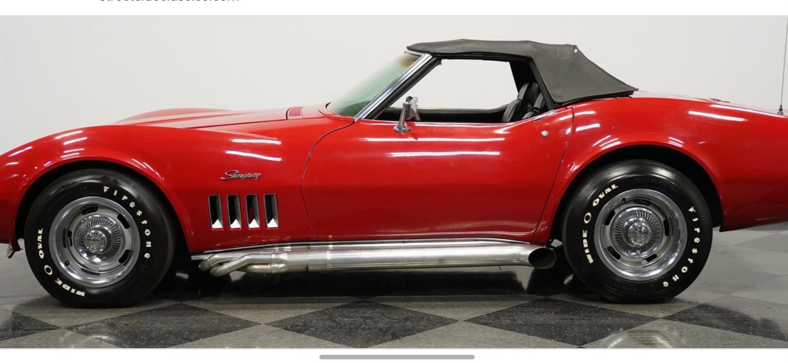 Red convertible sports car with black soft top displayed in showroom with chrome wheels and side exhaust pipe.
