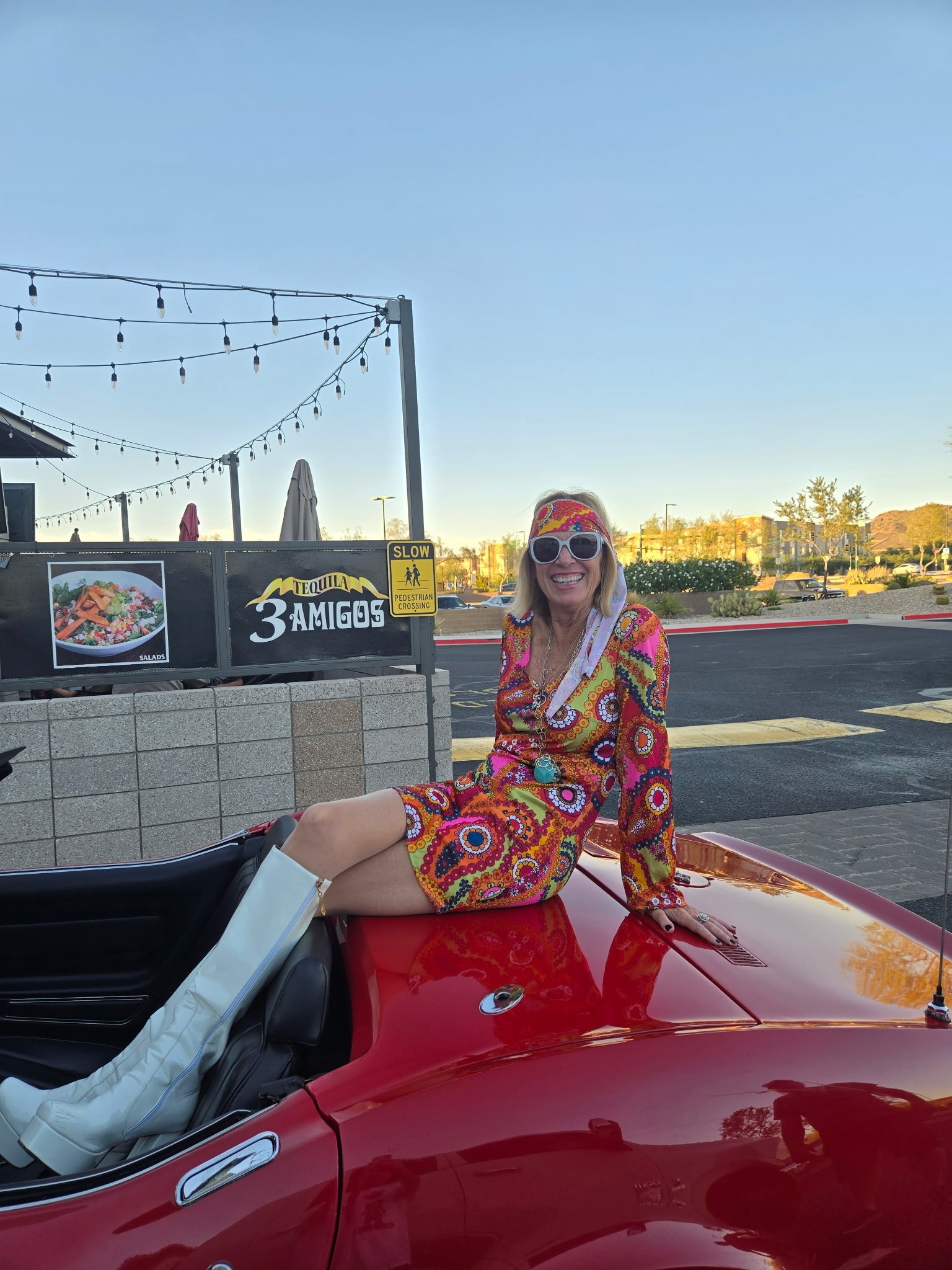 Woman in colorful patterned jacket and sunglasses posing on red convertible car at outdoor venue with string lights.