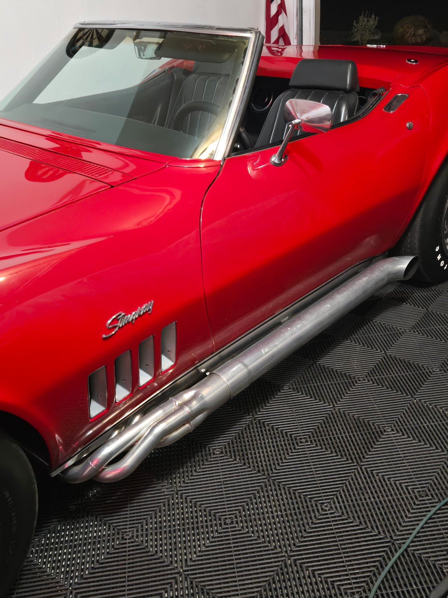 Bright red convertible sports car with chrome side pipes and black interior visible, displayed in garage setting.