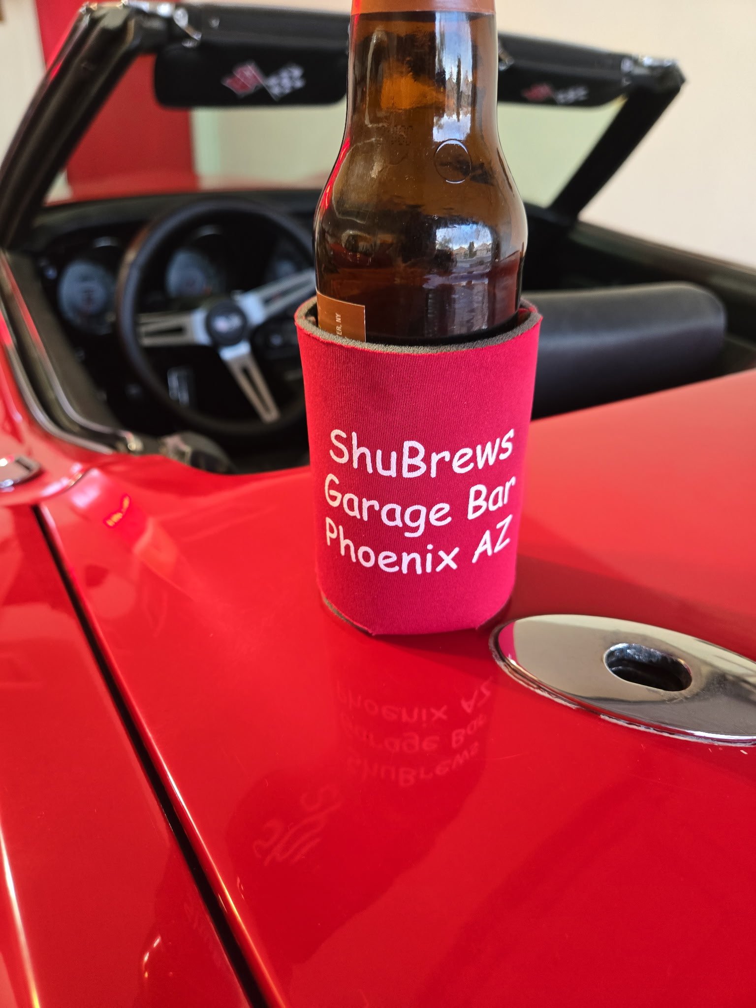 Beer bottle with ShuBrews Garage Bar Phoenix AZ koozie on red convertible car hood.